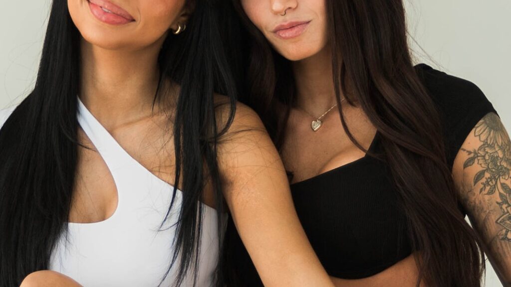 Two women with long dark hair, one in a white top and the other in black, stand close together. Featuring a tattooed arm and heart-shaped necklace, they embody inspiring real stories of patient results after a mommy makeover.