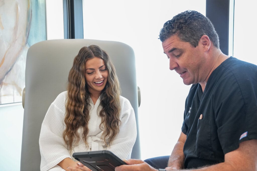 A woman in a white robe sits in a chair, smiling while a man in black scrubs shows her something on a tablet—perhaps discussing her upcoming Mommy Makeover consultation at their Draper, Utah clinic.