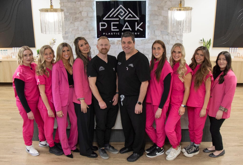 Ten staff members in pink medical scrubs and black professional attire standing before a Peak Plastic Surgery logo, ready to guide patients through the plastic surgery process.
