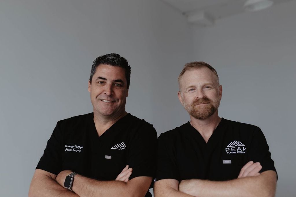 Two surgeons wearing black scrub tops with Peak Plastic Surgery branding, posing with their arms crossed.
