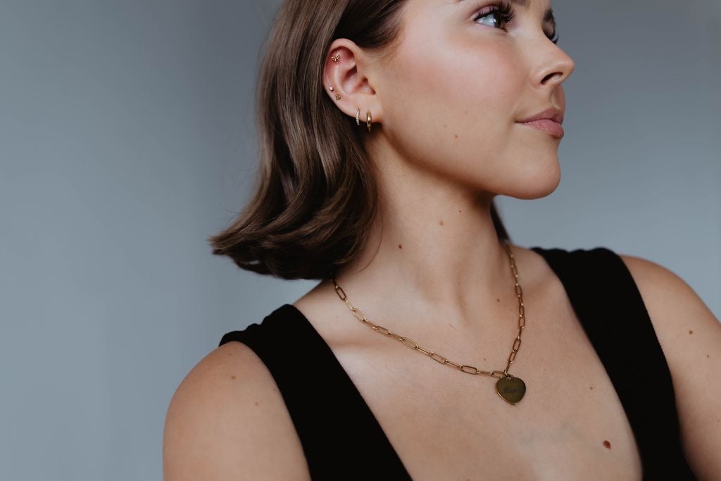 Woman with short brown hair wearing a black top, gold chain necklace with a pendant, and small gold hoop earrings, looking to the side against a neutral background. She appears radiant and refreshed, possibly following a neck lift in Draper, UT.