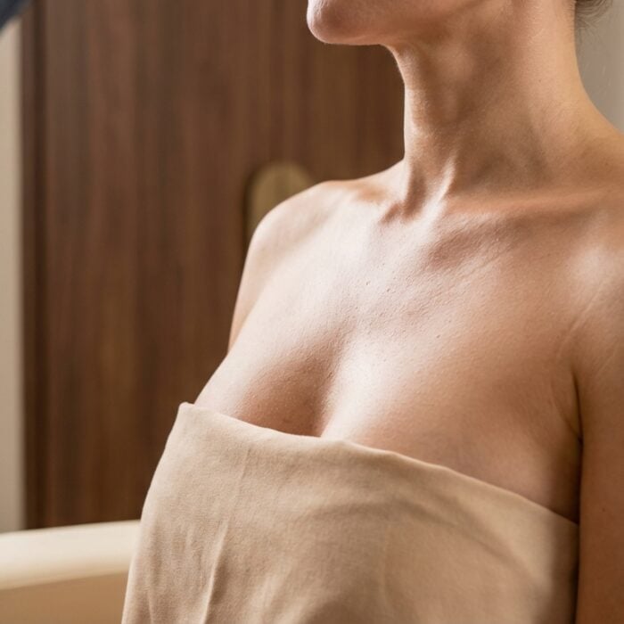 A woman seated indoors with a towel wrapped around her chest and shoulders, exposing her collarbone and upper chest.