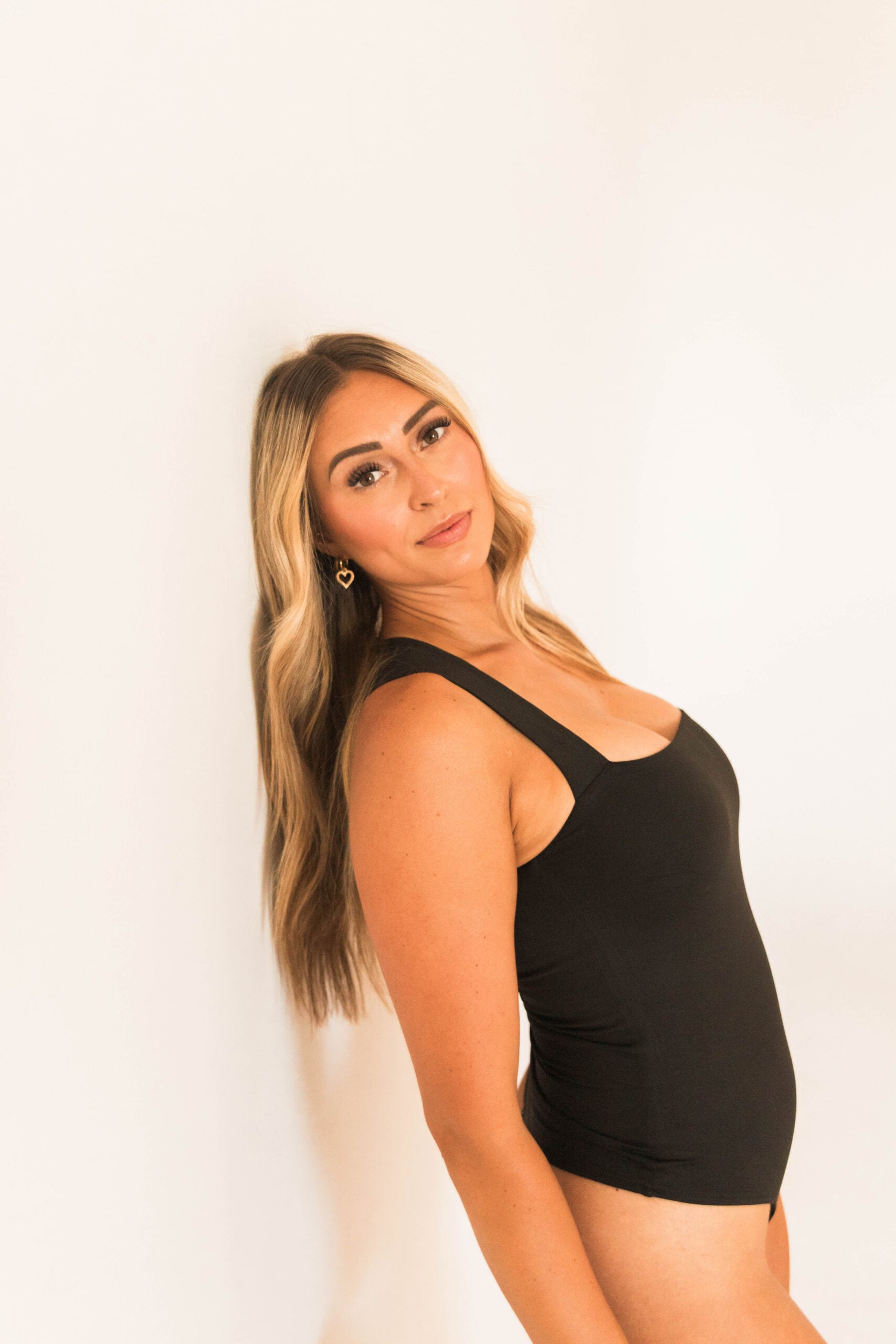 A woman with long, blonde hair wears a black tank top and stands against a plain white background, looking towards the camera.