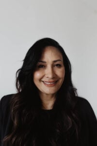 A portrait of a smiling woman with long, dark, wavy hair wearing a black top against a plain white background.