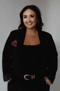 A woman smiling and wearing a black blazer with a pink “Peak Plastic Surgery” logo.