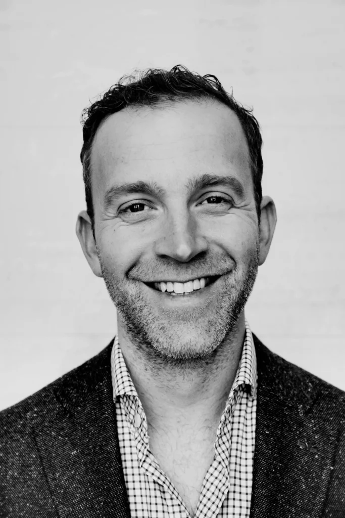 Dr. Griner, with short hair and light stubble, smiles at the camera. He is wearing a checked shirt and a dark blazer. The photo is black and white.