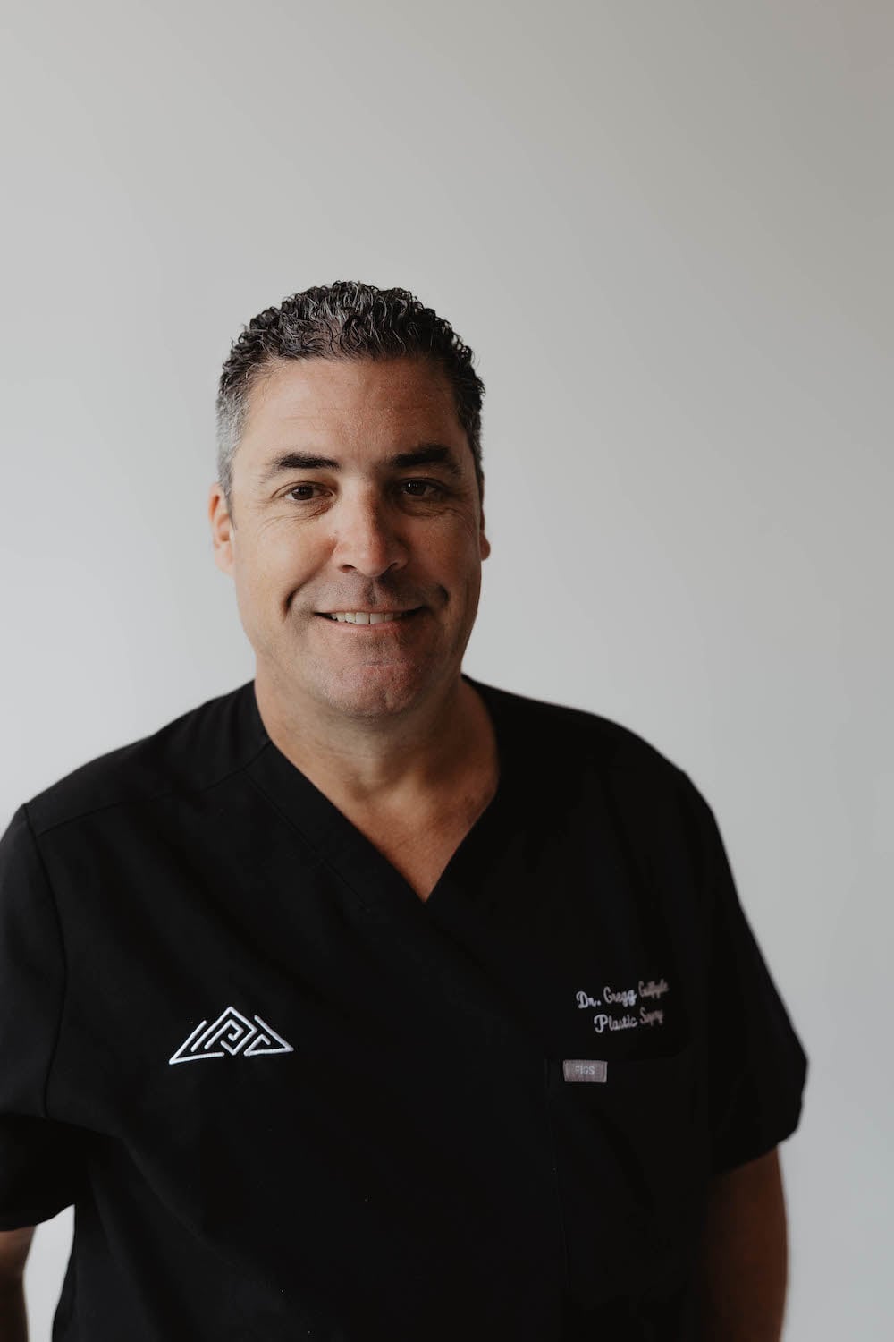 A man in a black medical scrub top stands against a plain, light background, smiling at the camera—an approachable and professional image reminiscent of Dr. Griner.