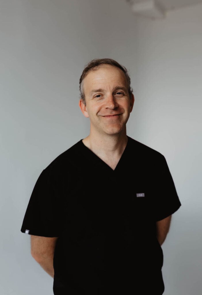 A man in black scrubs stands against a plain white background, smiling with his hands behind his back—much like Dr. Griner, known for his approachable professionalism and friendly demeanor.