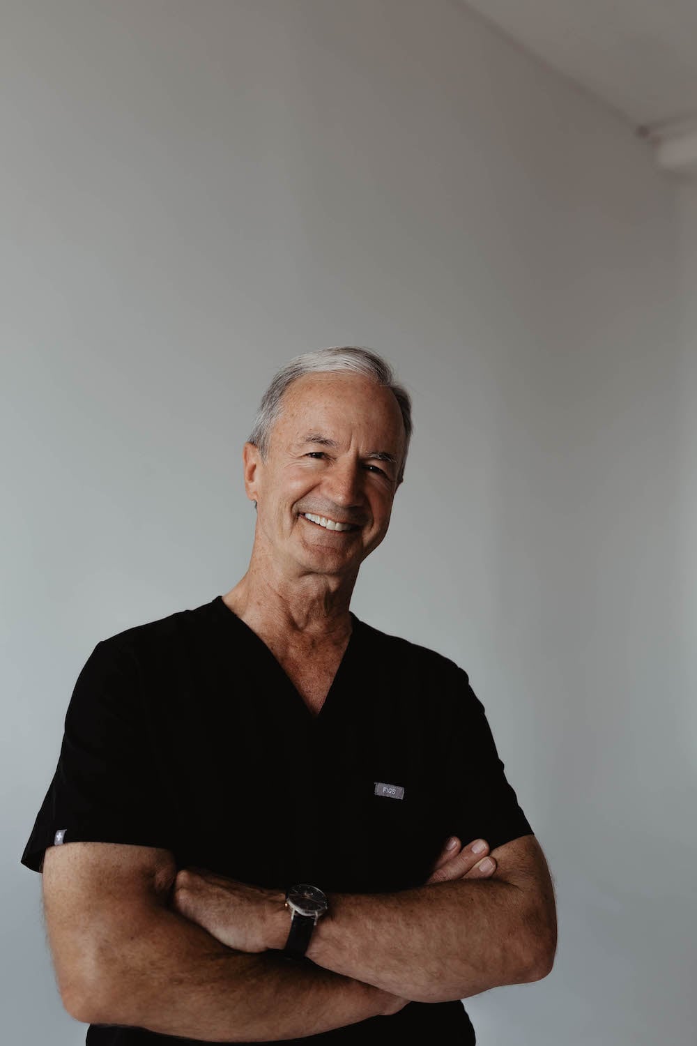 An older man with gray hair, wearing black scrubs, stands with arms crossed and smiles confidently against a plain white background—he could be Dr. Griner himself, radiating professionalism and warmth.