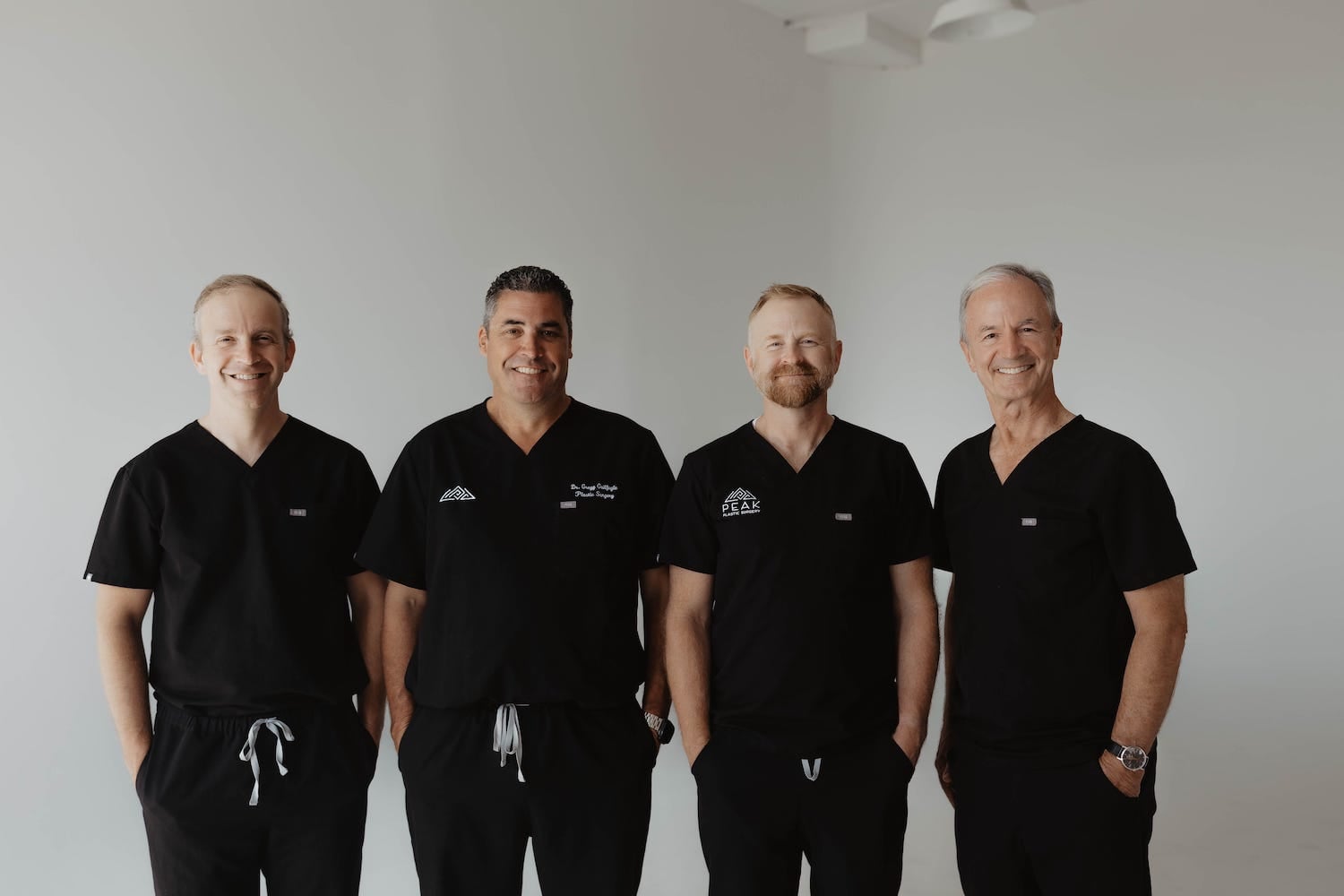 Four men in black medical scrubs stand side by side in a bright room with a white background, smiling at the camera.