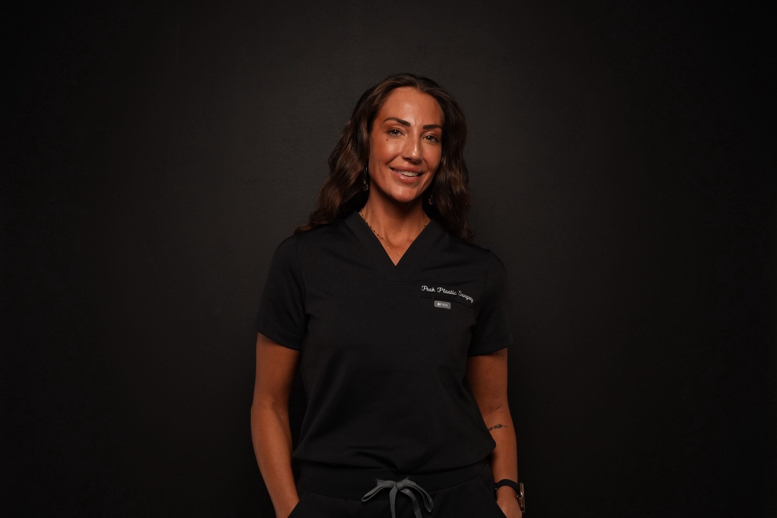 A woman wearing a black surgical scrub top with “Peak Plastic Surgery” embroidered on the chest.