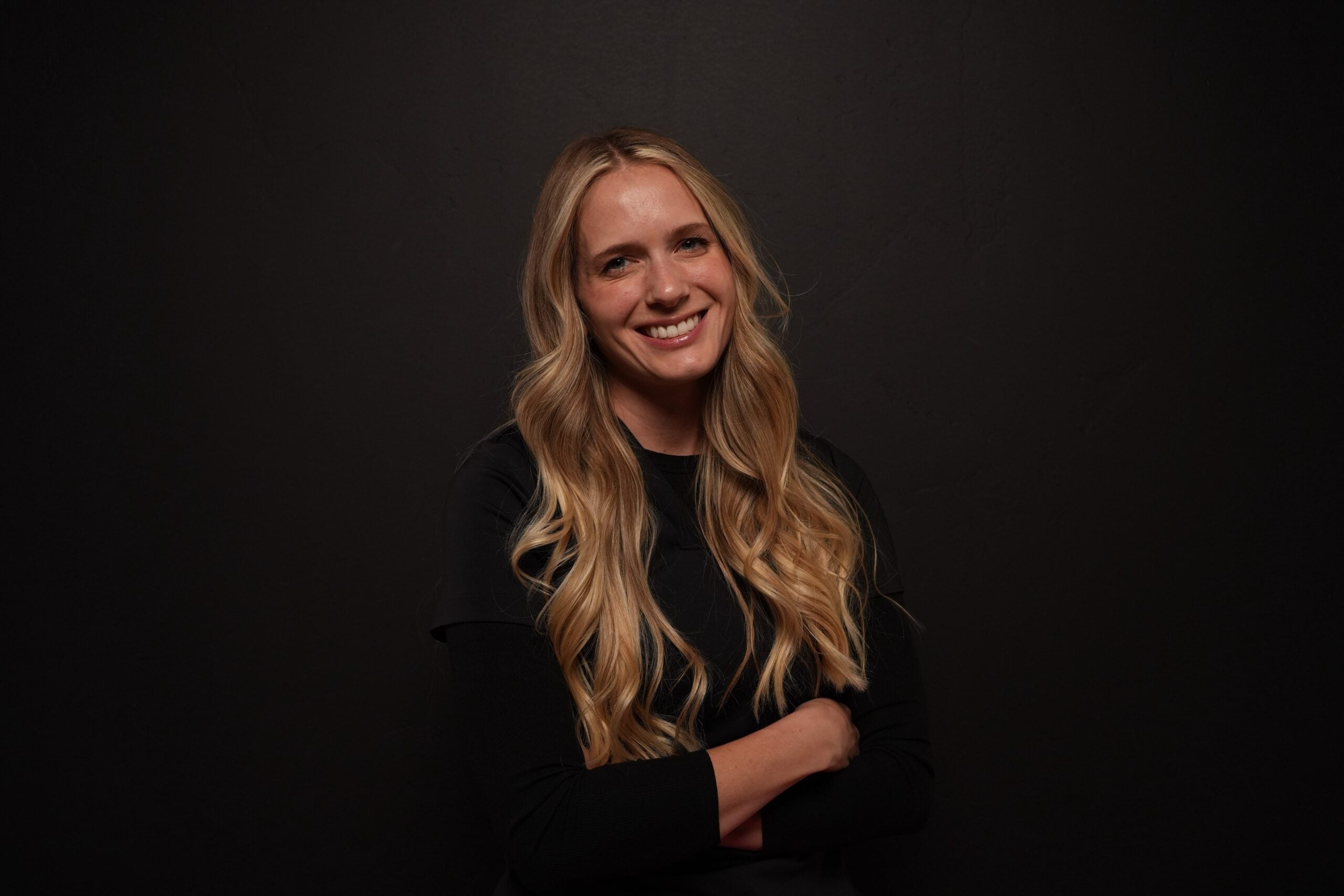 A smiling woman with long blonde hair, wearing a black shirt with arms crossed, against a black background.