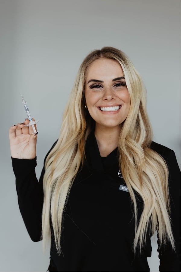 Woman with long blonde hair, wearing a black top, smiles and holds up a syringe against a plain, light background.