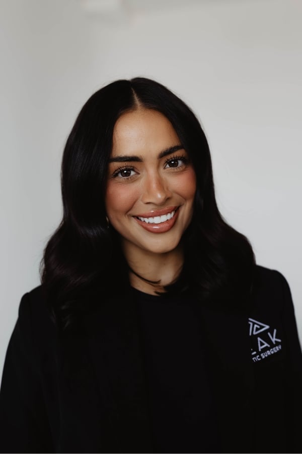 Woman with medium-length dark hair, wearing a black blazer and shirt, smiling at the camera against a plain light background.