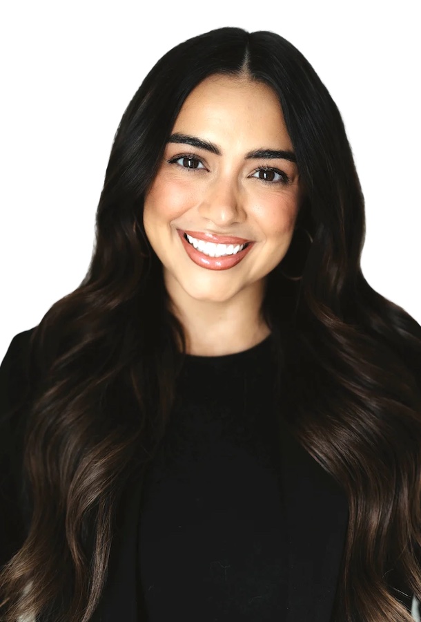 Smiling woman with long dark wavy hair, wearing a black top and black jacket, posed in front of a plain white background.