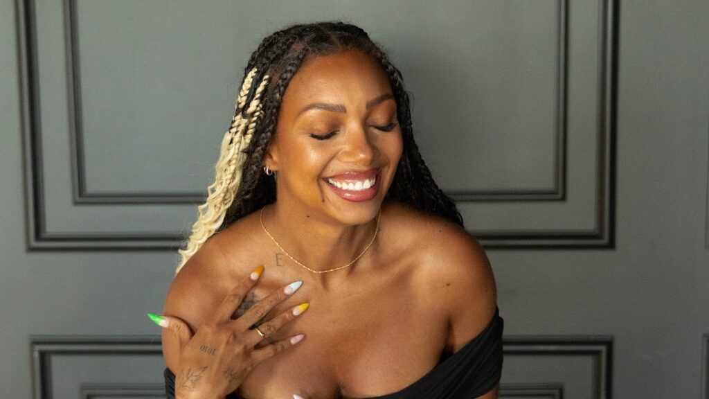 Woman with long braids and colorful nails smiles with eyes closed, embracing her Ideal Transformation in an off-the-shoulder top and gold necklace, posing confidently in front of a gray paneled wall—a true Mommy Makeover moment worthy of Myth-Busters.