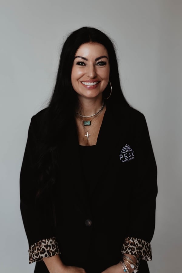 Woman with long dark hair, wearing a black blazer with a Peak Plastic Surgery logo, layered necklaces, and bracelets, standing in front of a plain background, smiling at the camera.