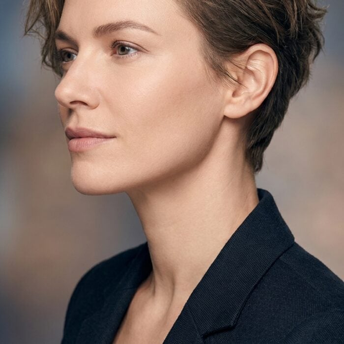 A person with short brown hair, wearing a dark blazer, poses in profile against a blurred, neutral background.