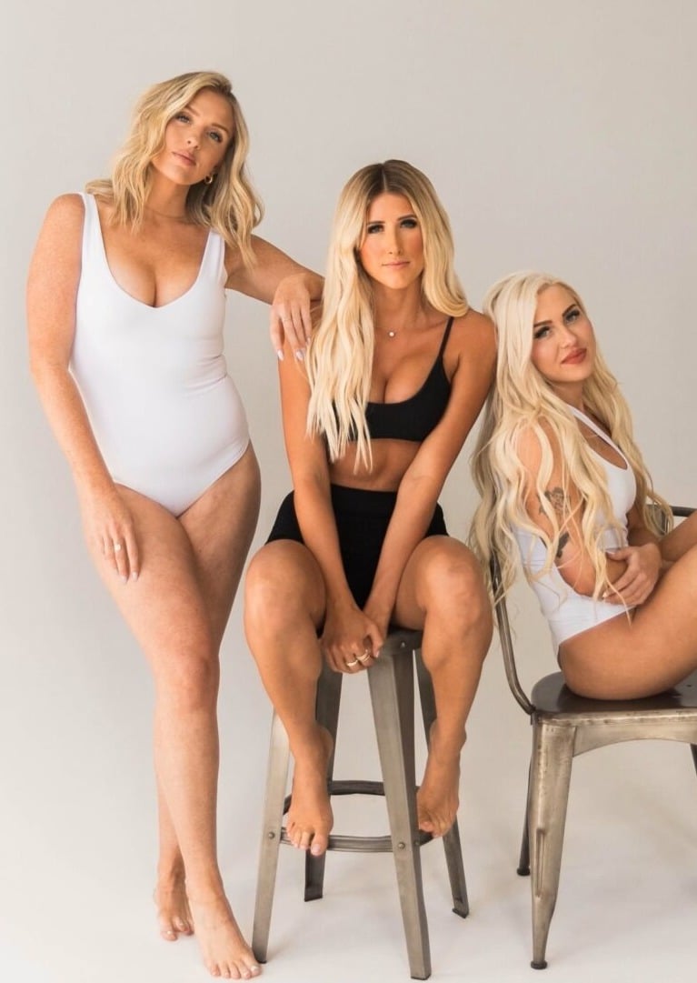 Three women with long blonde hair pose together against a plain background. One stands in a white bodysuit, one sits in black swimwear, and one perches on a stool in a white crop top and shorts.