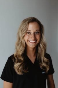 A woman with long, wavy blonde hair wearing a black shirt stands in front of a plain, light-colored background, smiling at the camera.