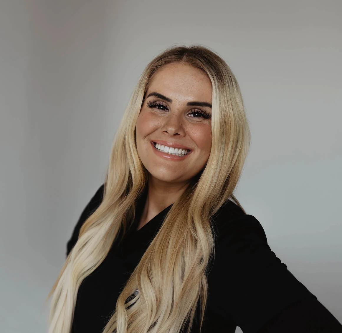 A smiling blonde woman wearing a black top against a plain light background.