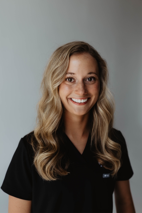 A woman with long, wavy blonde hair wearing a black shirt stands in front of a plain, light-colored background, smiling at the camera.