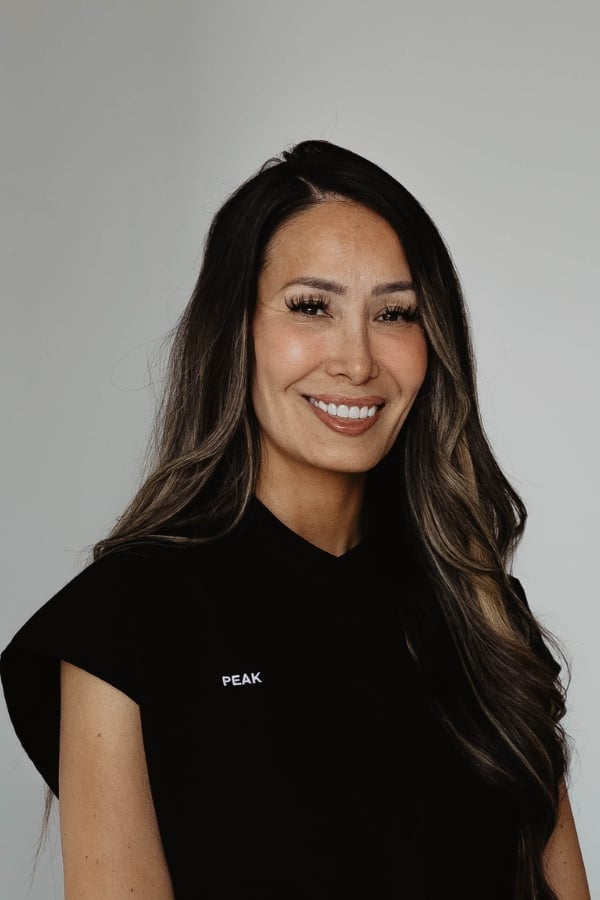 Woman with long brown hair smiles at the camera, wearing a black shirt with the word “PEAK” on it. Neutral gray background.