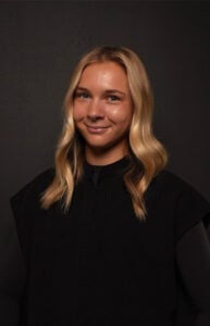 A smiling woman with blonde hair wearing a black shirt against a dark background.
