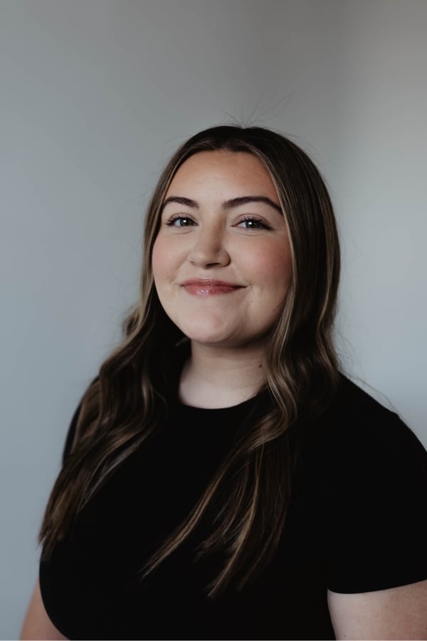 A young woman with long brown hair wearing a black shirt smiles slightly while looking at the camera against a plain light background.