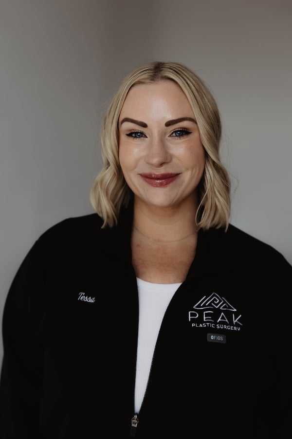 Woman with blonde hair in a black jacket that says “Tessa” and “Peak Plastic Surgery,” smiling at the camera against a plain background.